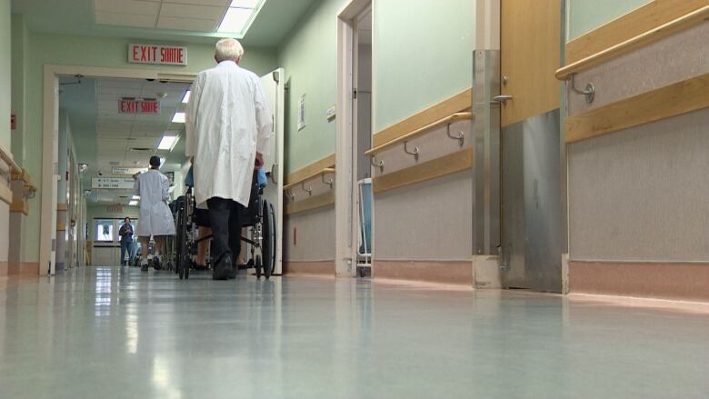 Two doctors in white lab coats walk down a hospital hallway toward another health-care provider standing at the end of the hallway; one of them pushing a patient in a wheelchair.