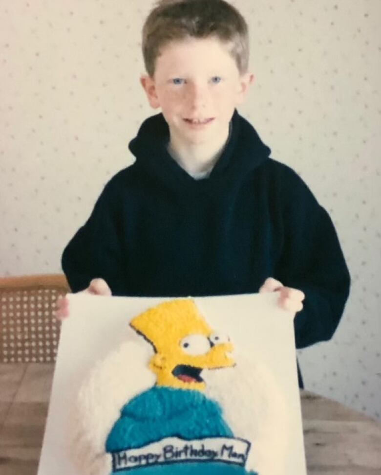 A boy in a black hoodie holds up a cake that has Bart Simpson on it.