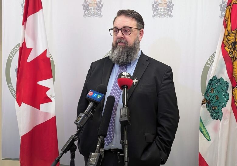 A man with a long beard and glasses, a purple tie and a dark suit speaks into microphones in a news conference room