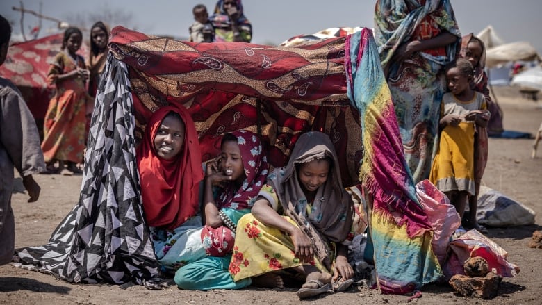 Three young girls in colourful dresses sit in the sand in makeshift tent, while other women and children stand nearby.