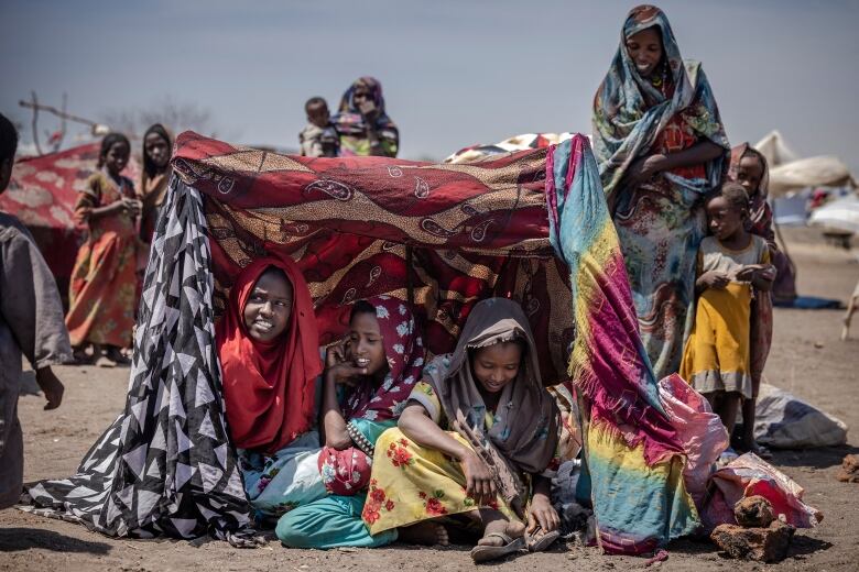 Three young girls in colourful dresses sit in the sand in makeshift tent, while other women and children stand nearby.