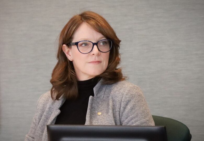 A woman with red hair and glasses wearing a grey jacket and sitting in front of a computer screen looks away.