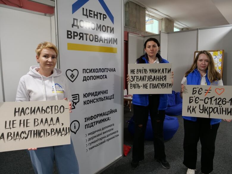 Three women stand holding hand-painted signs in Ukrainian.