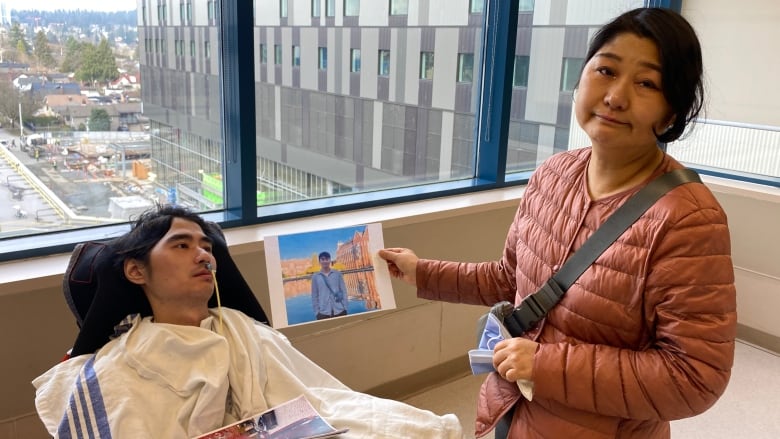 A woman stands next to a young man in a wheelchair. She holds a photo of him before he was in a vegetative state.