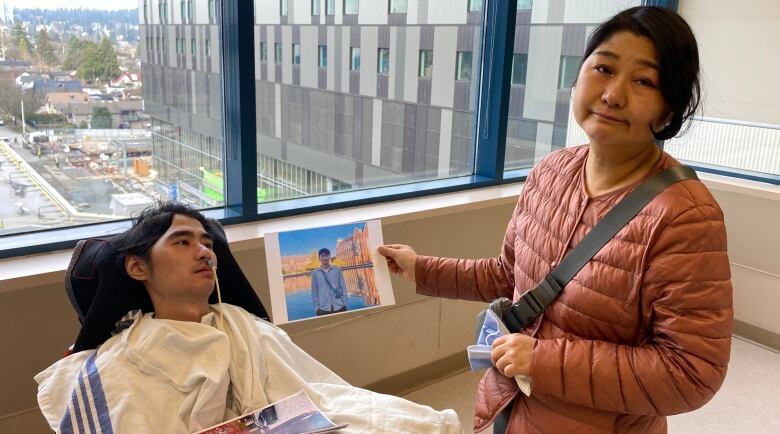 A woman stands next to a young man in a wheelchair. She holds a photo of him before he was in a vegetative state.