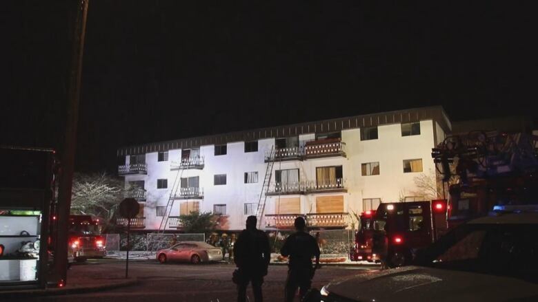 A three-storey white condo building is pictured with some windows borded up and some smahed. 