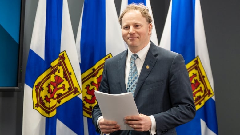 A man in a suit holds papers, standing in front of Nova Scotia flags.