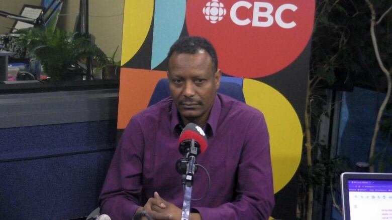 A man in a purple shirt sits at a microphone in a radio studio.