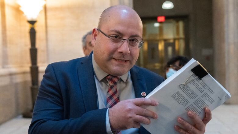 A bald man with glasses holds up a stack of papers, standing in front of a microphone