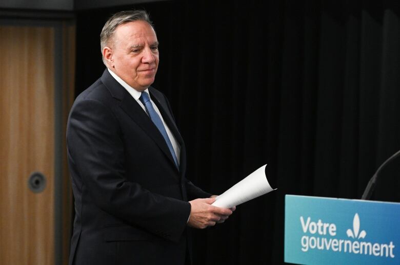 A white man in a suit holds papers as he walks up to a podium