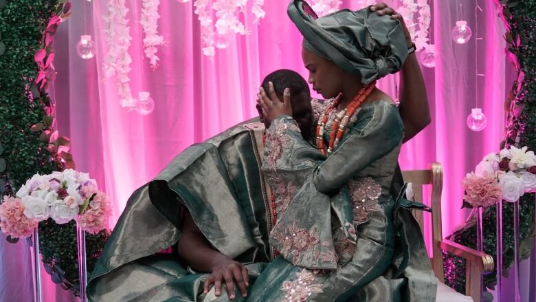 A man and woman, wearing elaborate green robes, embrace each other, heads close to one another. Behind them is bright pink light behind a light veil.