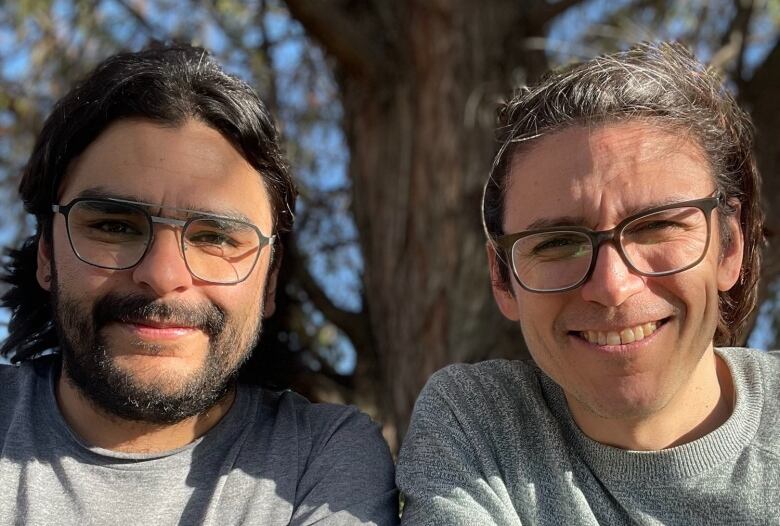 Two men, who are brothers, sit together at a table outside and smile for a photo.