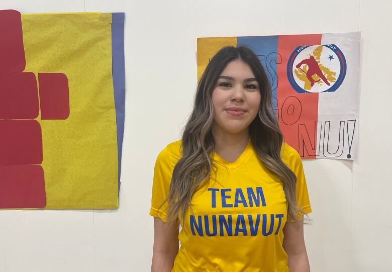 A young woman wearing a 'Team Nunavut' t-shirt stands in front of some posters on a wall.