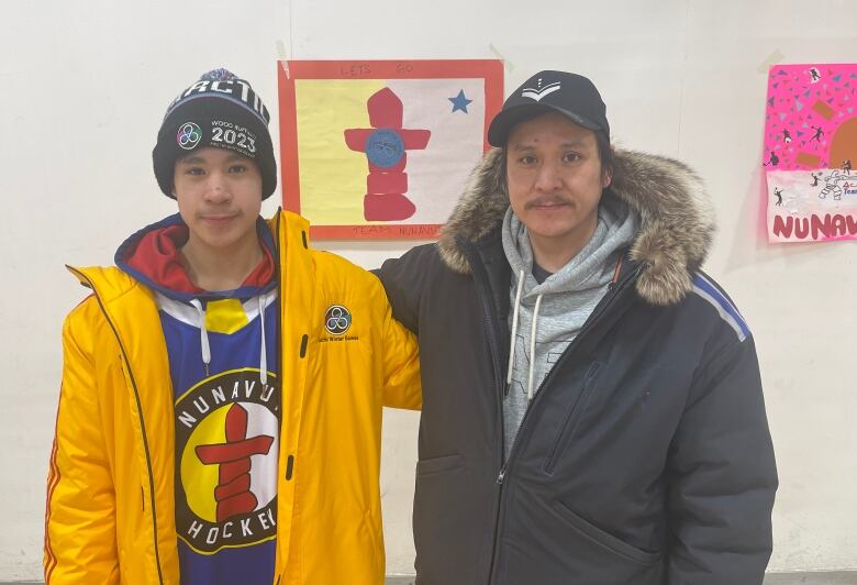 A man and a teenage boy stand with their arms around each other in front of some posters on a wall.