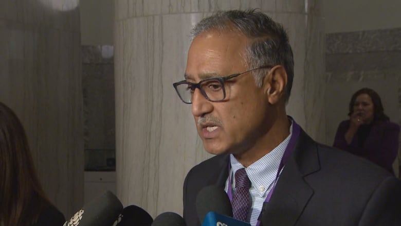 A man wearing a suit stands in a large building. He addresses a group of reporters.