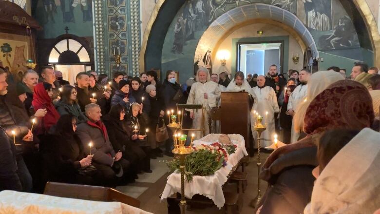 Mourners hold candles beside the open casket of a deceased man, with several people in religious garments shown.