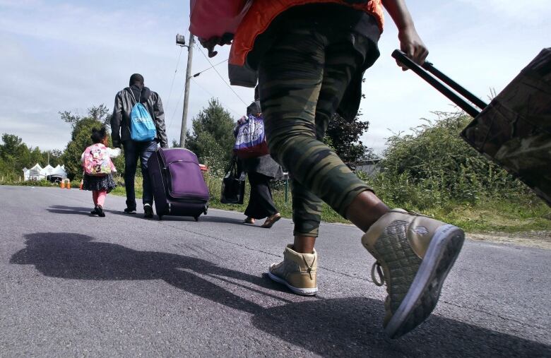 A family pulling suitcases is seen closeup from behind walking down a road. 