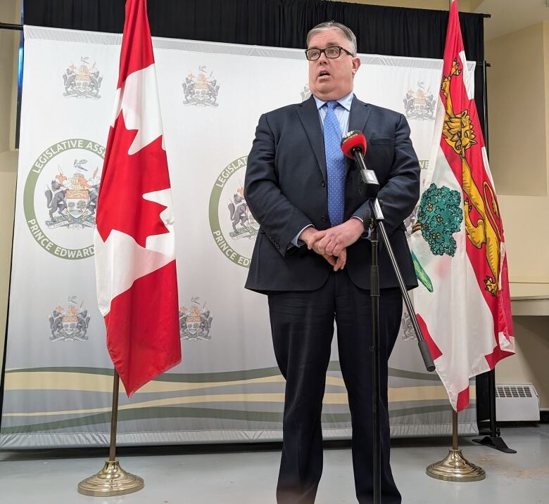 A man with glasses, a dark blue suit and light blue tie speaks into microphones in front of P.E.I. and Canada flags. 