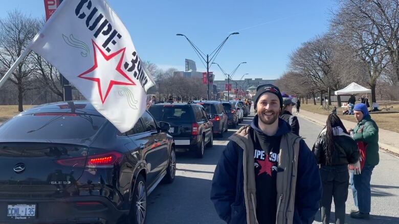 A young man in a toque smiles for the camera in front of a line of cars and a picket line. A CUPE flag waves beside him