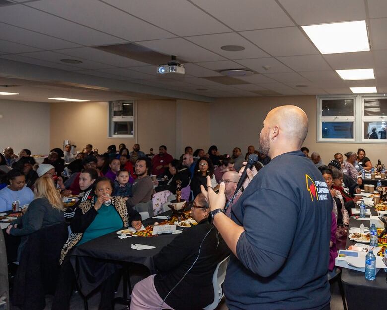 A bald black man with a microphone addresses a room full of people.