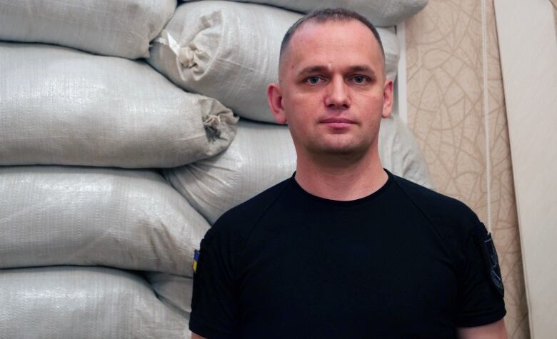 A man in a black T-shirt stands in front of a pile of sandbags.