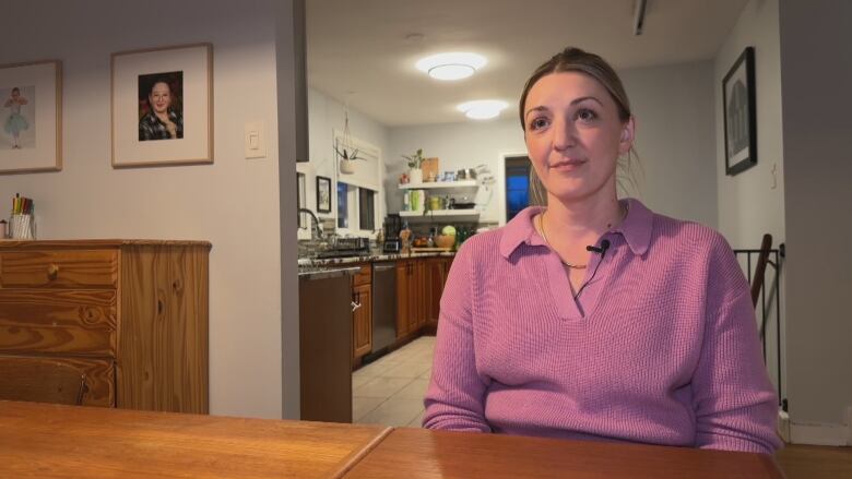 A woman sits at a dining room table with a kitchen in the background.