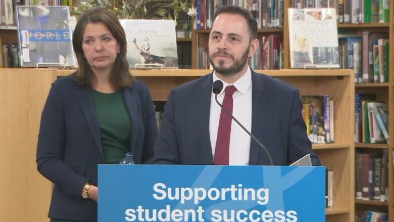 Education Minister Demetrios Nicolaides stands at a podium. Premier Danielle Smith stands beside him. They both look at an unpictured person who is asking them a question during a news conference in Calgary Friday.  