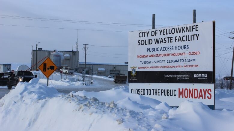 A sign on the right with information about Yellowknife's dump, surrounded by deep snow. A road, a truck and a building can be seen in the distance on the left. 