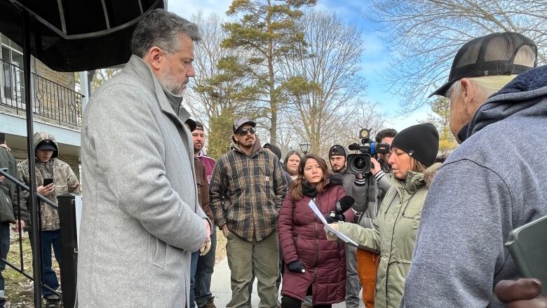 A Kanien'keh:ka woman in a coat reads a letter to a white man wearing a grey jacket with a crowd and TV journalists.