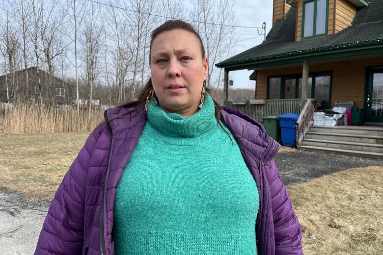 a woman wearing a turquoise sweater and purple jacket stands in a frontyard