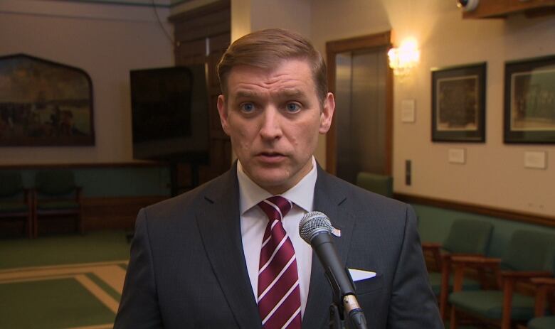 A man wearing a grey suit with a red tie stands behind a microphone in a lobby. 