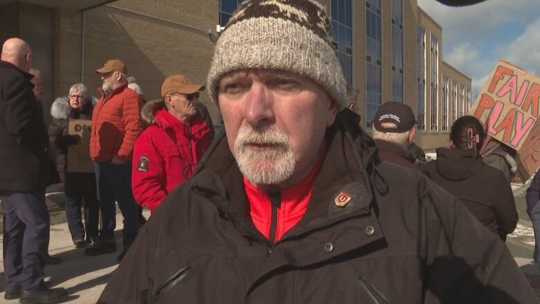 A man wearing a winter hat stands on the steps of Confederation Building in front of protestors. 