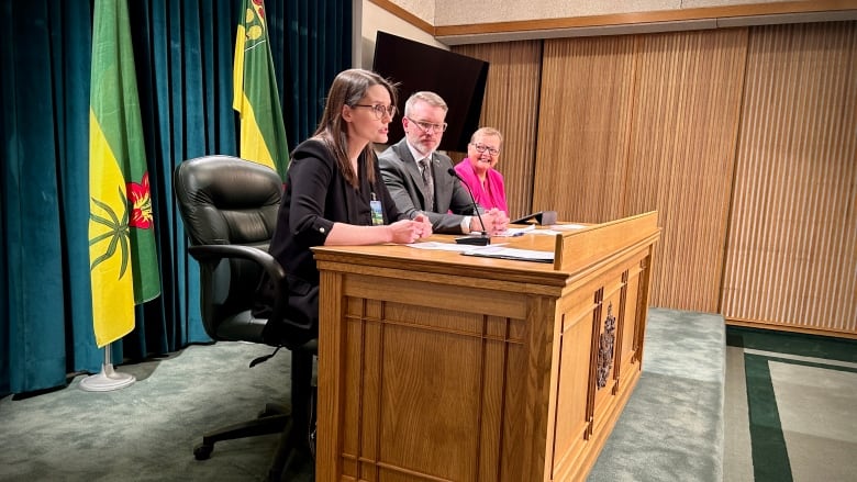 A woman in a black shirt, a man in a grey suit and a woman in a pink blazer sit at a wooden desk. 