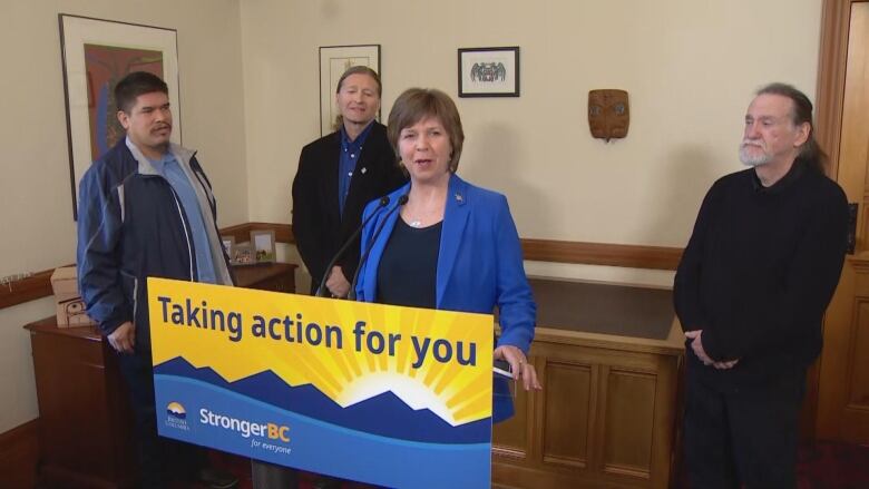 At a podium that read 'Taking action for you' a woman is speaking into the mic wearing a blue suit. Behind her are two men on the left, one wearing a black suit and the other wearing a blue jacket, and a man on the right wearing a black shirt. 