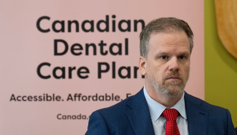 A man in a blue suit and red tie stands in front of a sign that reads 