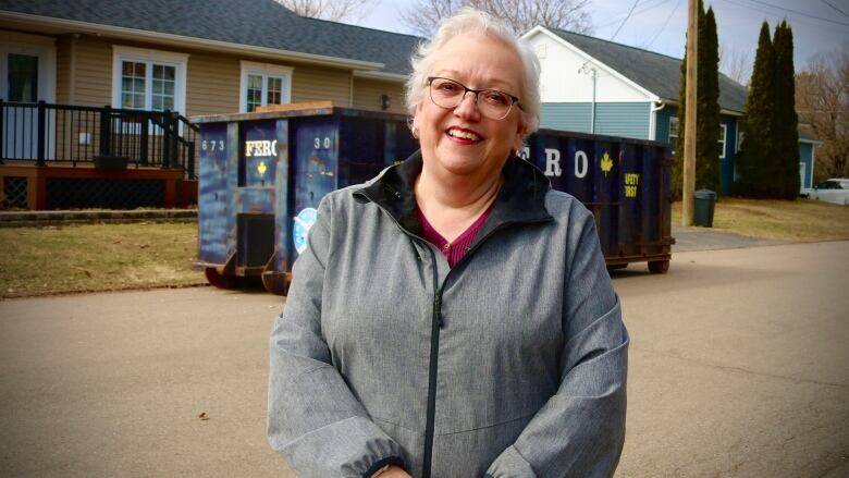  A smiling white-haired woman stands in front of a dumpster