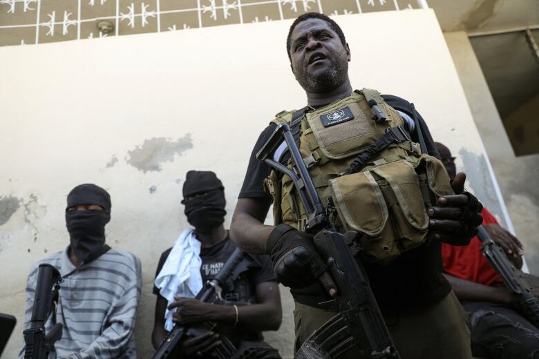 A gang leader holding a gun and wearing a combat vest stands surrounded by masked gang members.