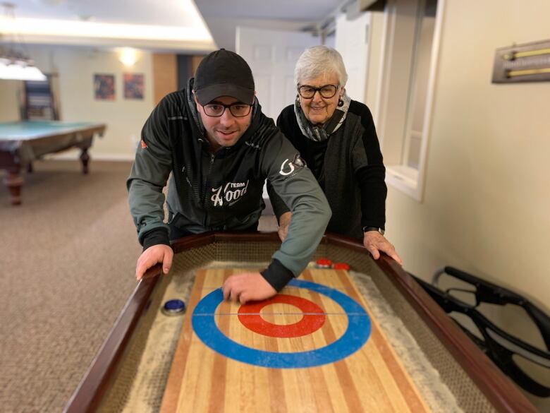 A young man wearing a black baseball cap and an older woman with white hair play shuffleboard.