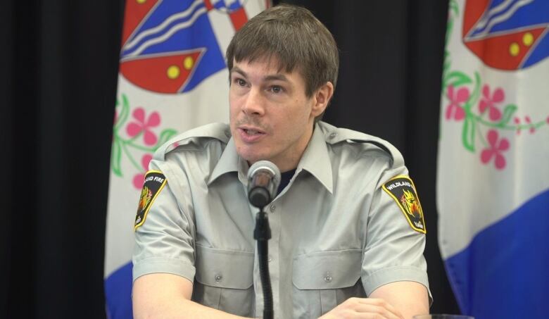 A man in a uniform shirt sits at a table speaking into a microphone.