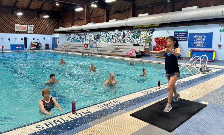 Tsay stands in a lunge position on a fitness mat in from of a swimming pool. Seven participants mimic her posture in the water.