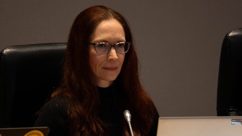 A woman in a black shirt with dark red hair and glasses sits in front of a long thin microphone