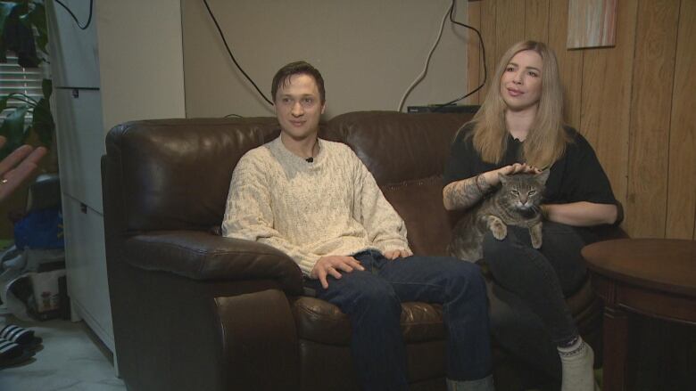 A man and a woman sit on a brown leather couch with a cat in the woman's lap. 