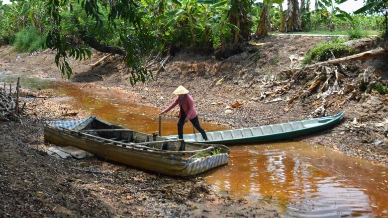 A person stands on a long boat that sits on a mostly dried up river bed. 