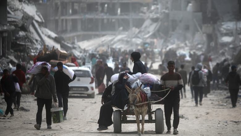 A cart led by an animal is shown at a distance on a dirt road. In the background are heavily damaged concrete buildings.