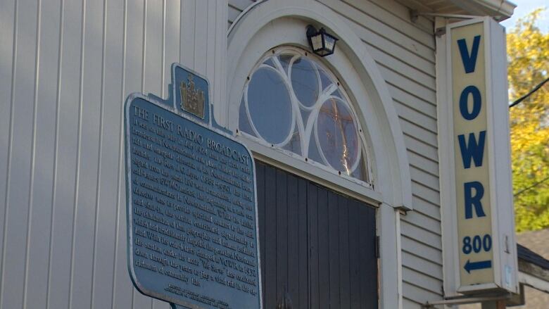 A large plaque is affixed to an outside wall of the church. Not all the words are legible but the top line of the sign reads 'The Very First Radio Broadcast.' Another sign nearby says 'VOWR 800' with an arrow pointing toward the church. 