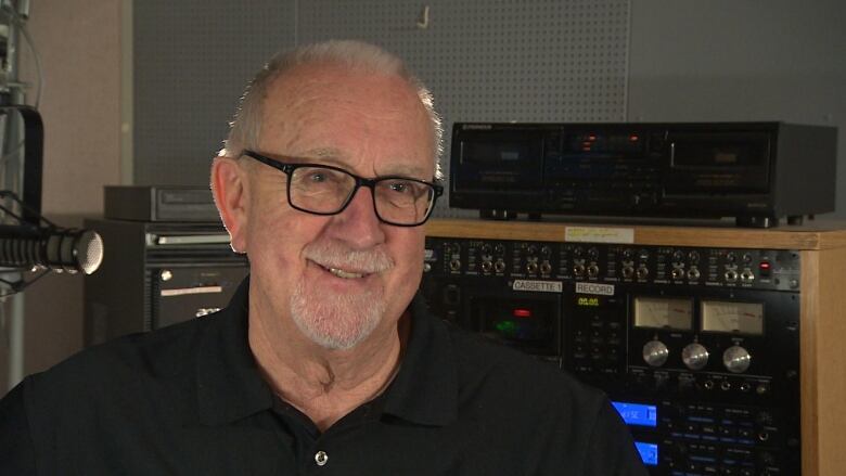 A man approximately in his 60's is smiling while sitting in a radio booth. A mic and a variety of radio equipment surround him.