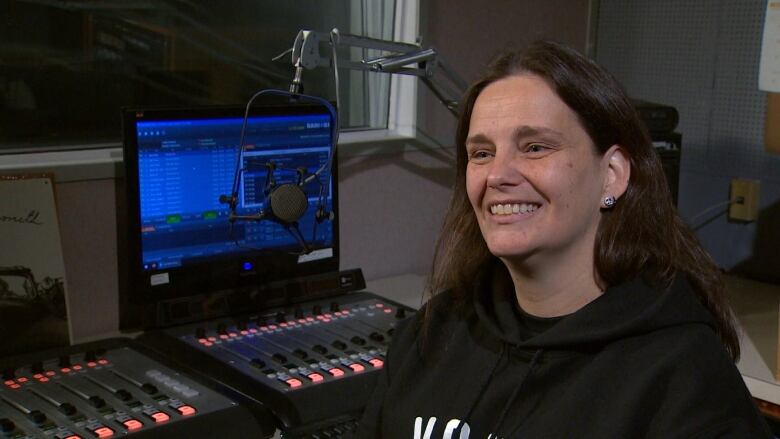 A woman smiles in front of a microphone and a radio soundboard as well as a computer screen. 