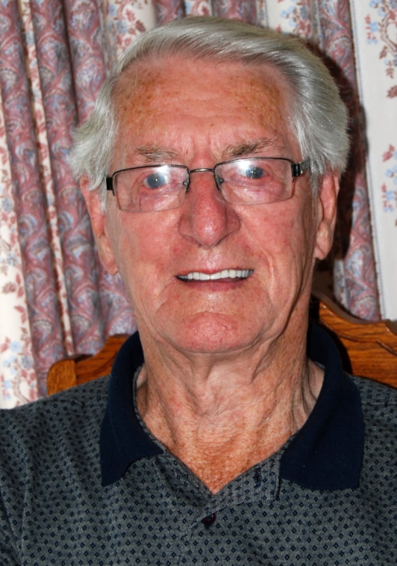 An old man smiles in front of a paisley-patterned curtain.