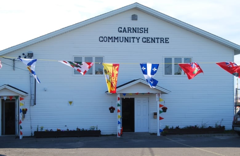 A small A-frame building has the words 'Garnish Community Centre' inscribed over the door. Several provincial Canadian flags are hanging on a banner in the foreground.