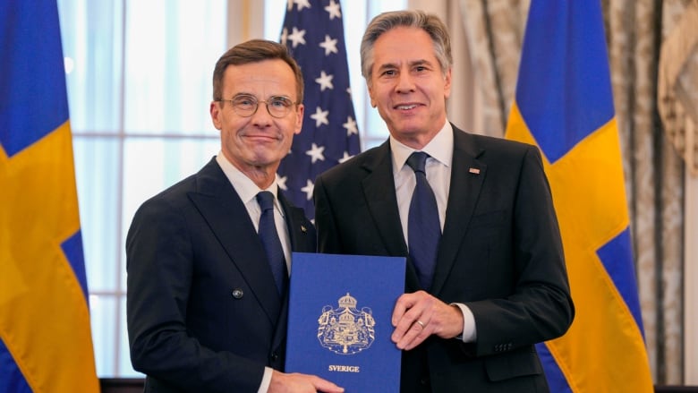 Two cleanshaven men wearing suit and tie, one of them bespectacled, pose for a photo while holding a leather-bound book together.